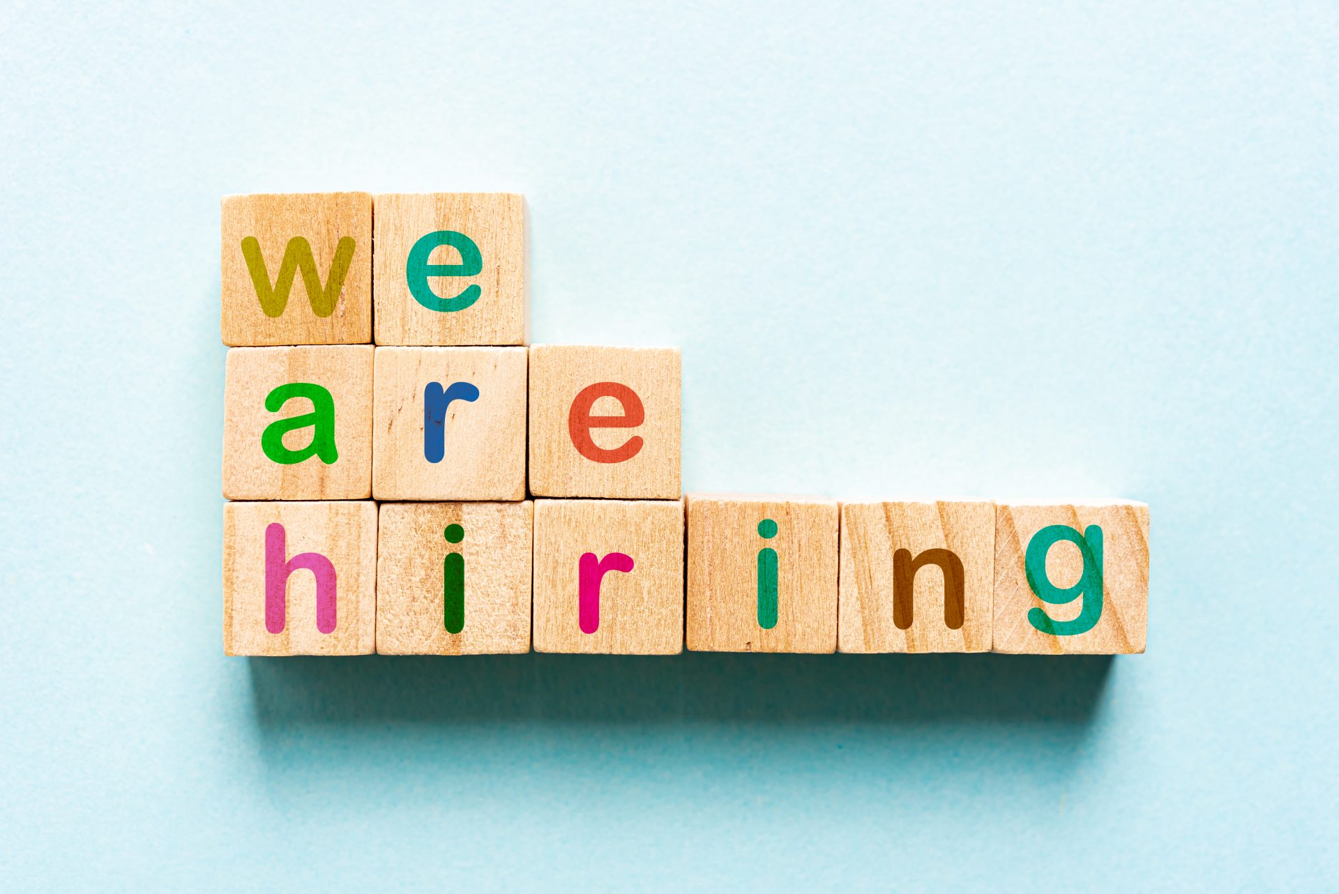 iStock-1206312395 Wooden letter blocks arranged to spell out 'We are hiring,' with colorful letters on a light blue background. The playful design emphasizes a welcoming and positive job recruitment message.