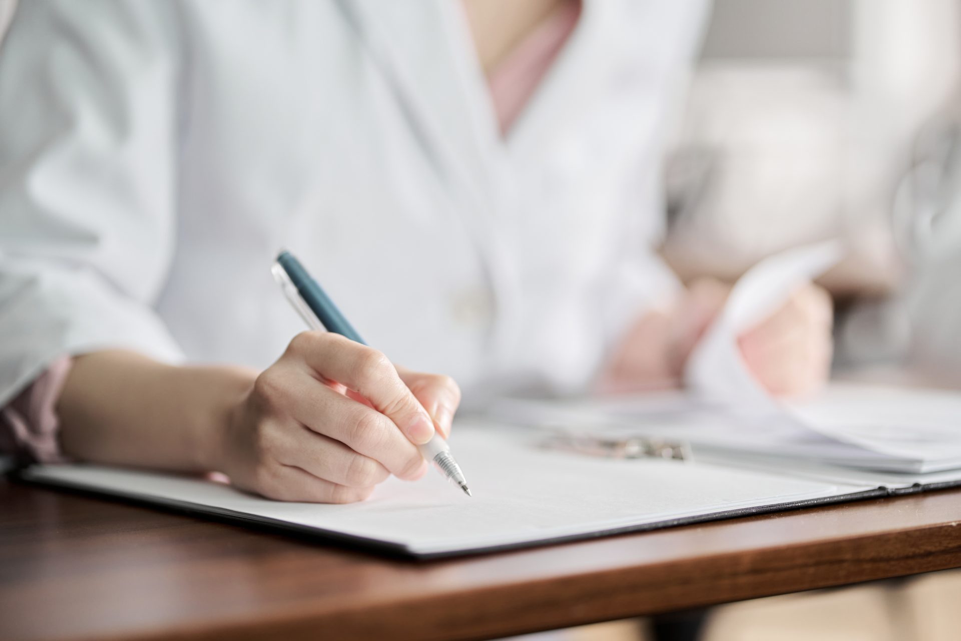 Close-up of a healthcare professional's hand holding a pen while writing on a clipboard. The individual is wearing a white coat, with documents and a blurred background indicating a clinical or office setting.