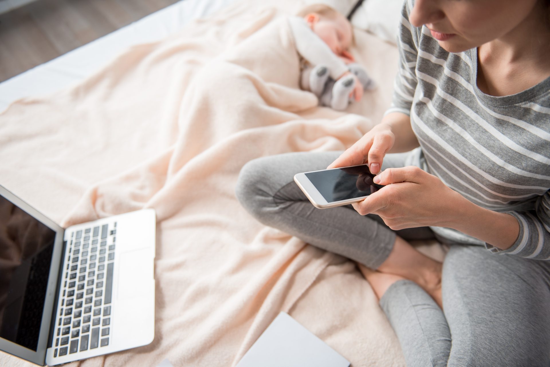 iStock-900588462 A mother in a striped shirt and gray leggings sitting on a bed, using her smartphone while a laptop rests nearby. In the background, her baby sleeps peacefully under a soft beige blanket, creating a serene and multitasking moment.